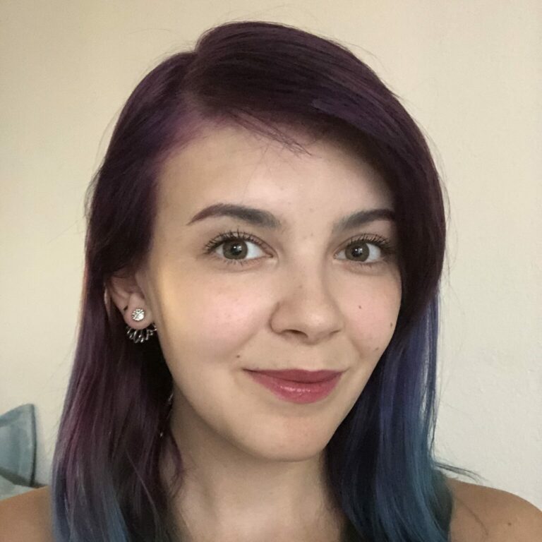 A selfie of Amber Williams. She has long hair in a side part which is dyed an ombre of dark purple and blue. She's smiling in the photo and has basic makeup and floral earrings on.